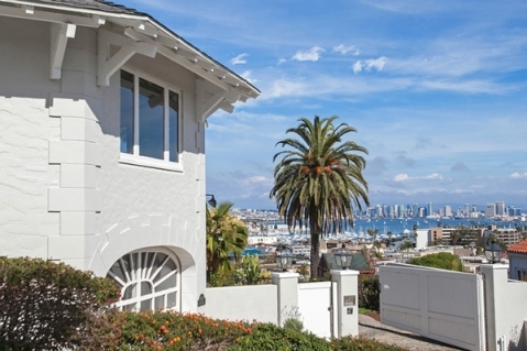 Light-colored house at an elevation with large windows and an arched feature, looking out over a city skyline, a bay filled with boats, and palm trees under a bright blue sky.